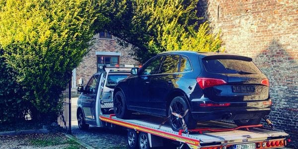 Zwarte Audi Q5 vastgesjord op het kantelende laadplatform van de takelwagen voor een bakstenen woning in helder zonlicht