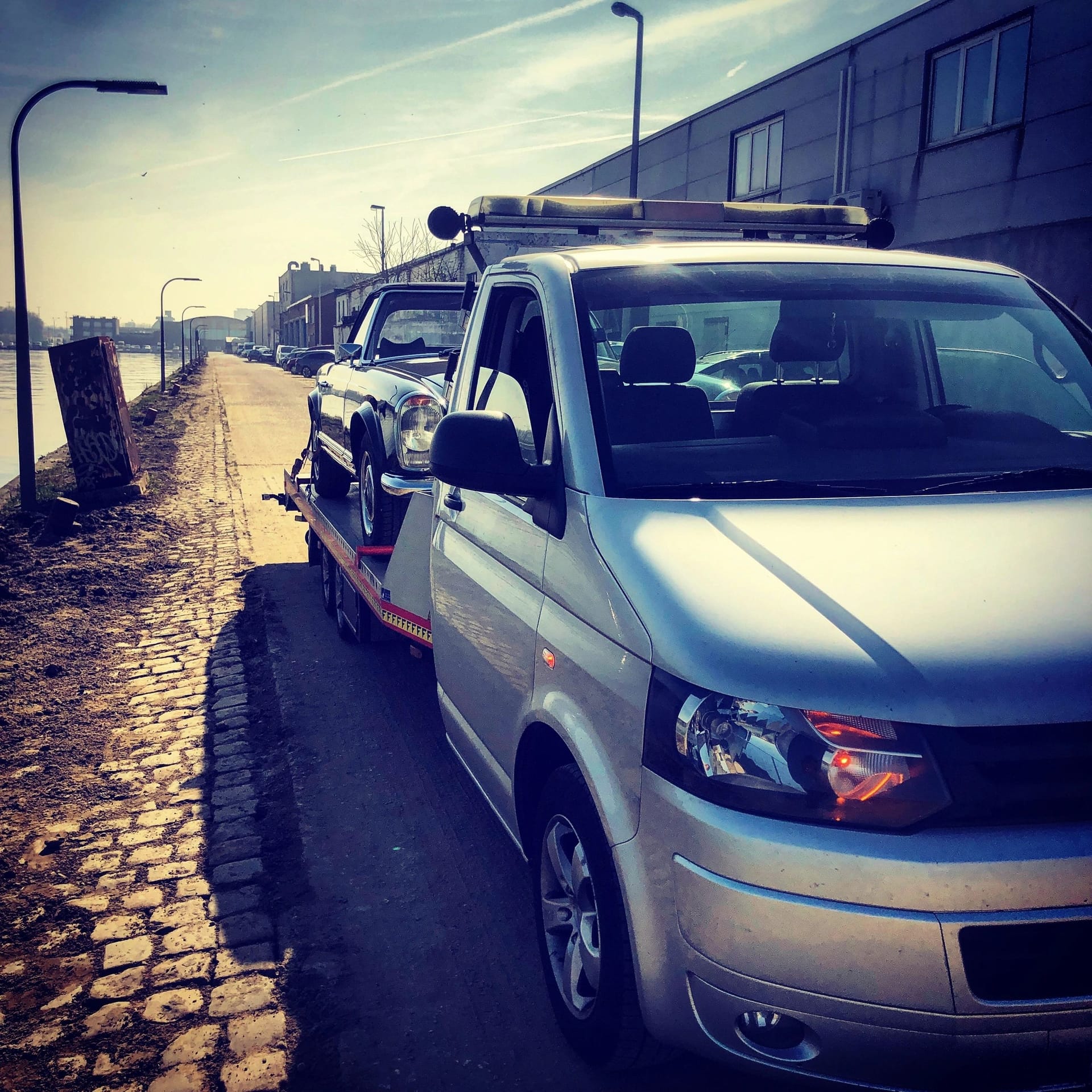 Zilvergrijze VW Transporter takelwagen met een klassieke wagen op het laadplatform, rijdend langs een kade bij het water in warm avondlicht