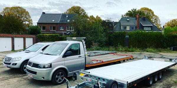 Zilvergrijze takelwagen met leeg laadplatform en aanhangwagen naast een Mercedes SUV in een residentiele buurt